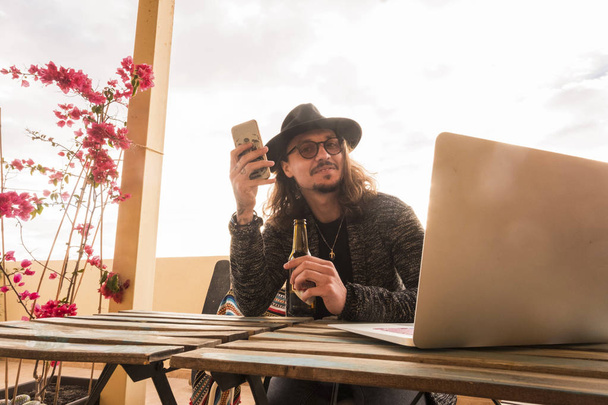 trendy man with hat and eyeglasses drink a beer while use the phone to communicate with friends and people. laptop on the table outdoor terrace to work free - Photo, Image