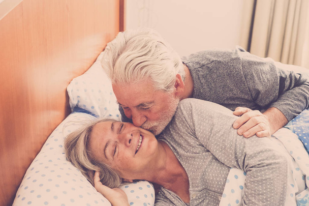 Couple of senior man and woman waking up and smiling with a hug while are in the bed at home. Vintage filter and light in the back. The man kiss the woman with love - Foto, afbeelding