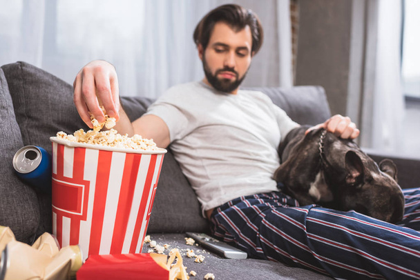 handsome loner sitting with bulldog and taking popcorn on sofa in living room  - Fotografie, Obrázek