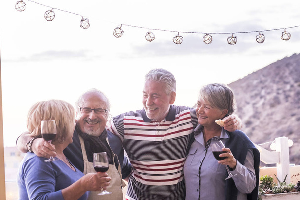 group of 4 seniors, two couples, stay togheter and have fun enjoing a cup of wine outdoor in the rooftop. vacation elderly concept and friendship. sunlight anche clear sky - Photo, Image