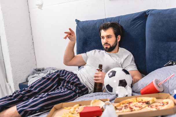 красивый одиночка смотрит футбол с пивом в спальне и бросает салфетку
 - Фото, изображение