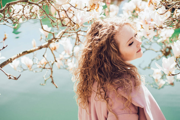 curly young lady in a pink coat posing next to a blooming magnolia in a park in the rays of the sun, spring - Фото, изображение