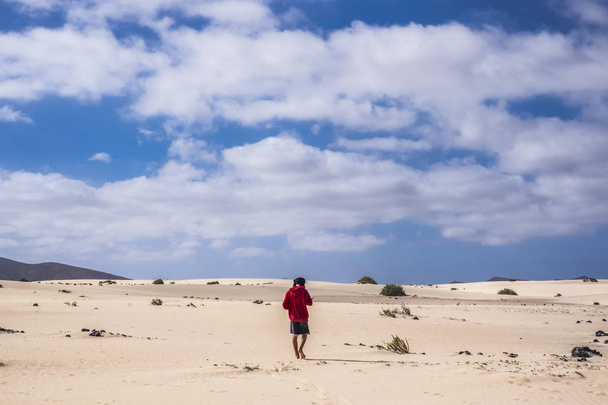 lonely young man with red jacket walking in the niddle of the desert in fuerteventura canary islands spain. nude feet like lost with mountains and nowhere around him. blue sky and yellow sand. - Fotografie, Obrázek