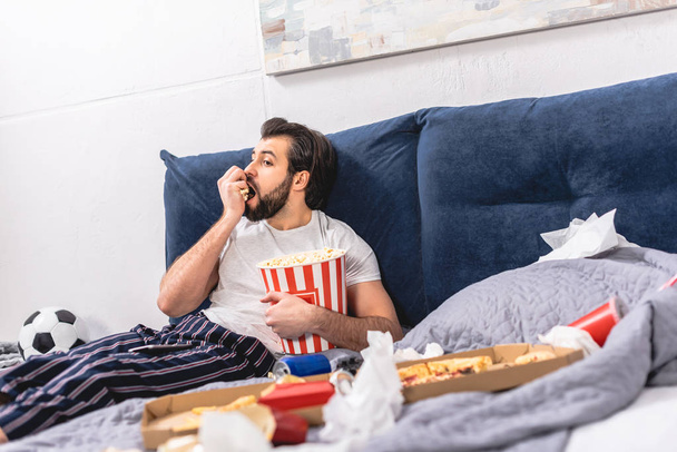 handsome loner eating popcorn and watching tv in bedroom - Foto, afbeelding