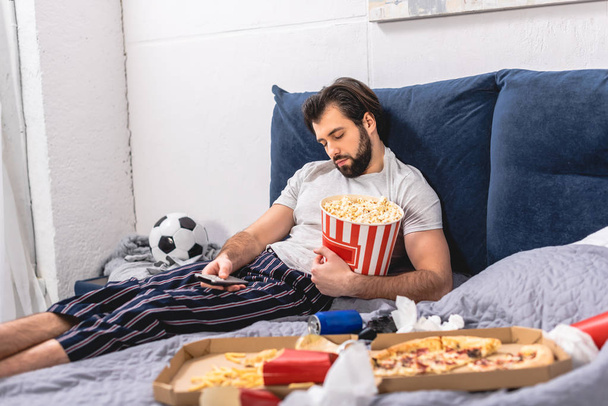 handsome loner sleeping with popcorn and remote control in bedroom - Photo, Image