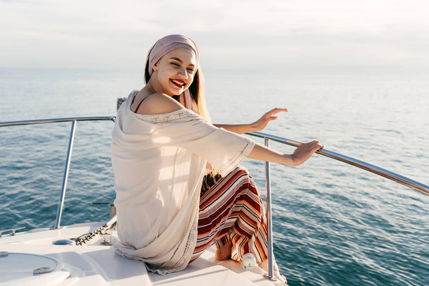Gelukkig rijk meisje in stijlvolle zomer kleding zit op haar witte jacht in de zon en de glimlach - Foto, afbeelding