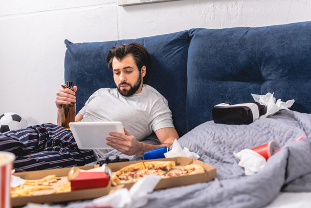 guapo solitario usando tableta en la cama en el dormitorio y sosteniendo botella de cerveza
 - Foto, Imagen