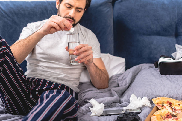 loner having headache with hangover and putting pill into glass of water in bedroom - Photo, Image