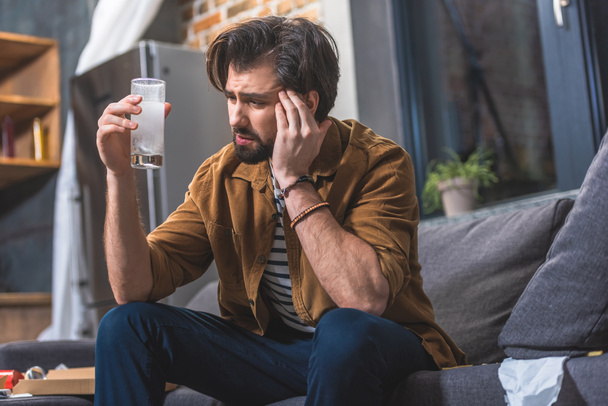 loner having headache and hangover and looking at glass of water with pill at living room - Photo, Image