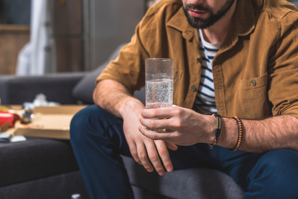cropped image of loner with hangover holding glass of water at living room - Fotografie, Obrázek