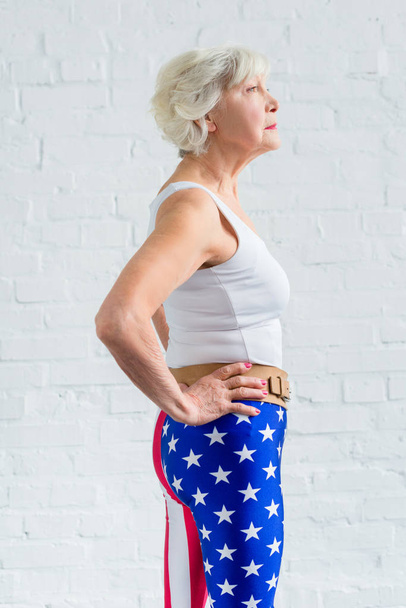 vista lateral da mulher idosa em sportswear de pé com as mãos na cintura e olhando para longe
 - Foto, Imagem