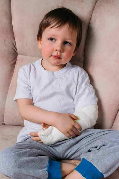 a small boy is sitting on a chair with a broken arm, in a cast. hospital. medicine. danger. pain. to break limbs. violence. - 写真・画像