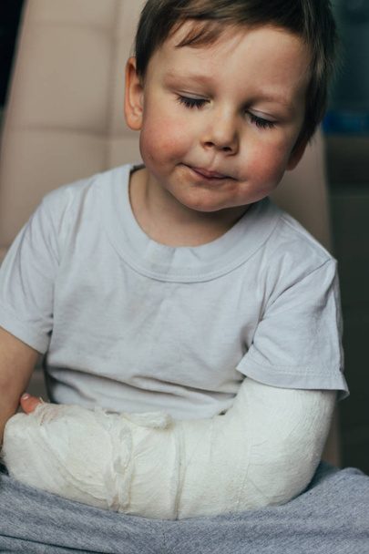 a small boy is sitting on a chair with a broken arm, in a cast. hospital. medicine. danger. pain. to break limbs. violence. - Фото, изображение