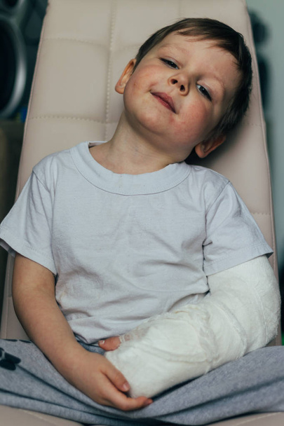 a small boy is sitting on a chair with a broken arm, in a cast. hospital. medicine. danger. pain. to break limbs. violence. - Foto, imagen