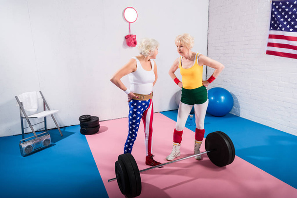 Hochwinkelaufnahme von älteren Sportlerinnen, die mit den Händen auf der Taille stehen und einander im Fitnessstudio anschauen - Foto, Bild