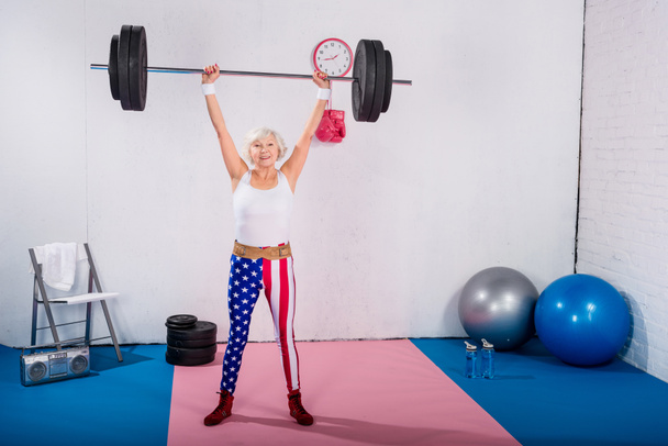 sportive senior woman lifting barbell and smiling at camera  - Fotografie, Obrázek