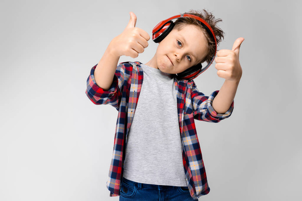 A handsome boy in a plaid shirt, gray shirt and jeans stands on a gray background. A boy in red headphones. The boy shows the class sign. - Фото, изображение