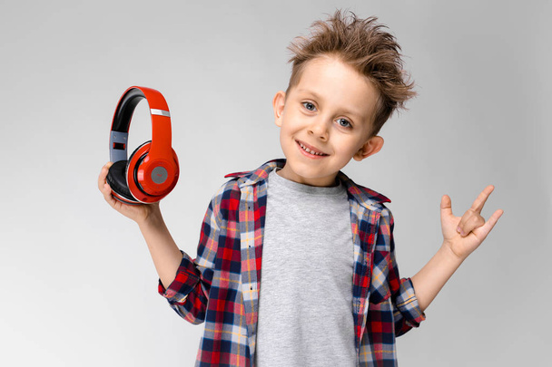 A handsome boy in a plaid shirt, gray shirt and jeans stands on a gray background. A boy in red headphones. The boy took off his headphones. - Zdjęcie, obraz