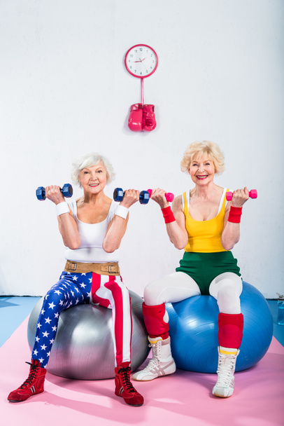 senior women in sportswear training with dumbbells while sitting on fitness balls and smiling at camera - Valokuva, kuva