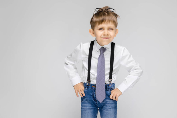 Ένα όμορφο αγόρι σε ένα λευκό πουκάμισο, ζαρτιέρες, γραβάτα και ελαφριά τζιν στέκεται σε γκρι φόντο. Γυαλιά στο κεφάλι - Φωτογραφία, εικόνα