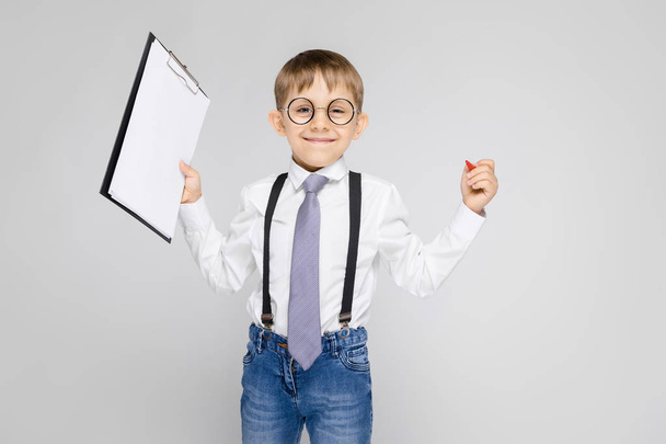 A charming boy in a white shirt, suspenders, a tie and light jeans stands on a gray background. The boy holds a pen and sheets for notes - Photo, Image