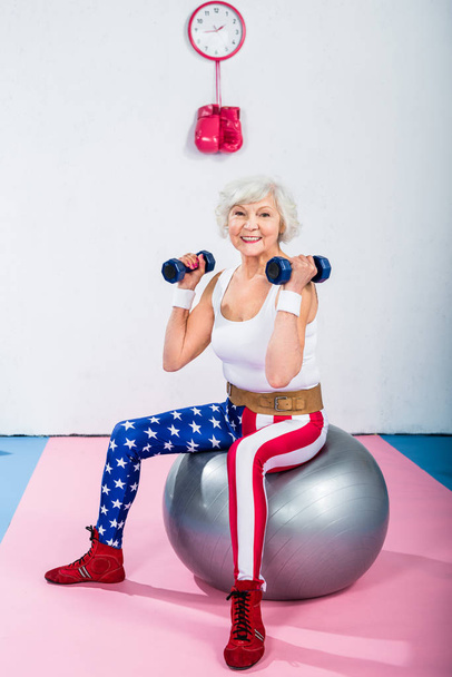 happy sporty senior woman with dumbbells sitting on fitness ball and smiling at camera - Фото, зображення