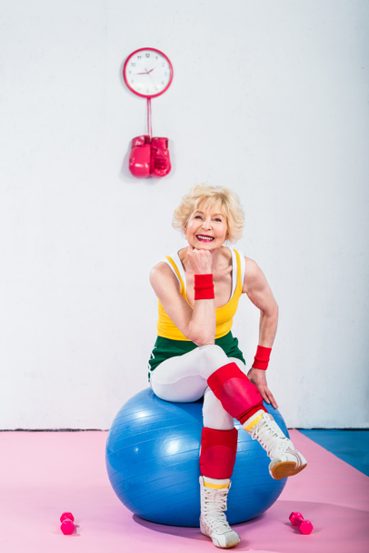 feliz deportivo senior mujer sentado en fitness pelota y sonriendo a la cámara
 - Foto, Imagen