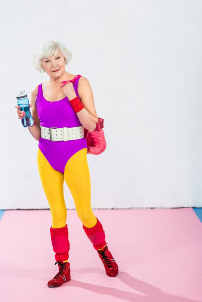 full length view of sportive senior lady with bottle of water and boxing gloves smiling at camera - Foto, imagen