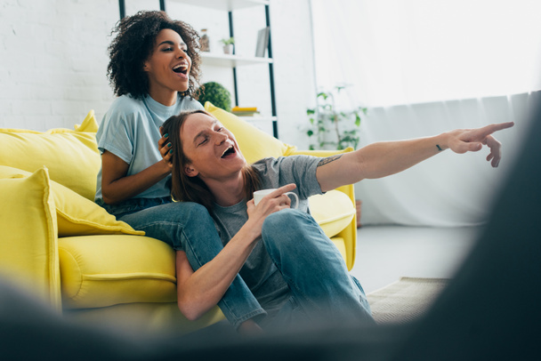laughing young man with coffee cup pointing on tv screen to smiling african american girlfriend  - Foto, imagen