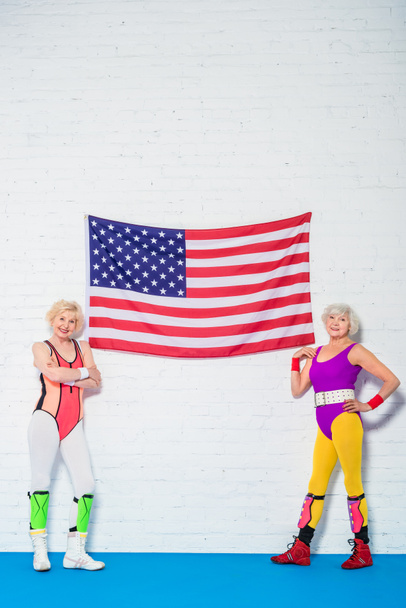 Ganzkörperansicht schöner älterer Sportlerinnen, die neben der amerikanischen Flagge stehen und in die Kamera lächeln   - Foto, Bild