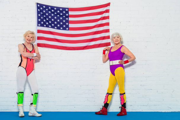 hermosas deportistas senior de pie cerca de la bandera americana y sonriendo a la cámara
   - Foto, Imagen