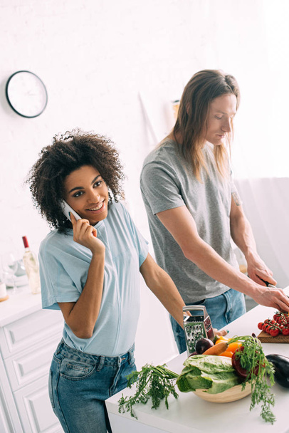 lächelnde Afroamerikanerin spricht auf Smartphone, während ihr Freund in der Küche Kirschtomaten schneidet  - Foto, Bild