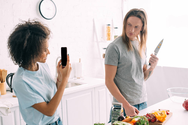 smiling african american woman shooting boyfriend posing with knife on smartphone - 写真・画像