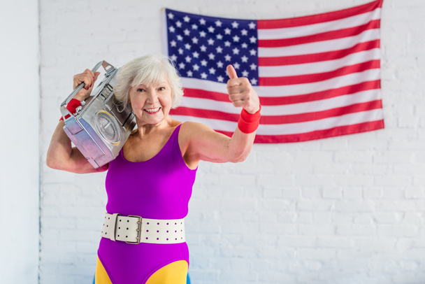 happy senior woman in sportswear holding tape recorder and showing thumb up - Photo, Image