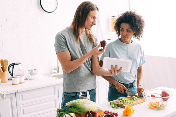 young man holding onion and showing recipe on digital tablet to girlfriend while she cutting salad leaves at kitchen - Foto, imagen