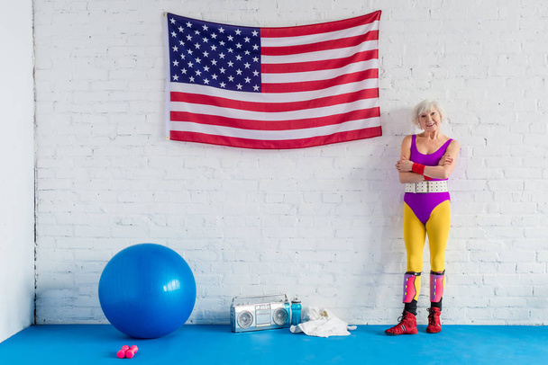 bela esportista sênior de pé com braços cruzados perto da bandeira americana e sorrindo para a câmera
 - Foto, Imagem