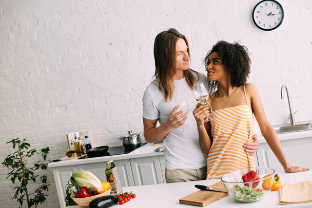 junges multiethnisches Paar mit Weißweingläsern in der Küche  - Foto, Bild
