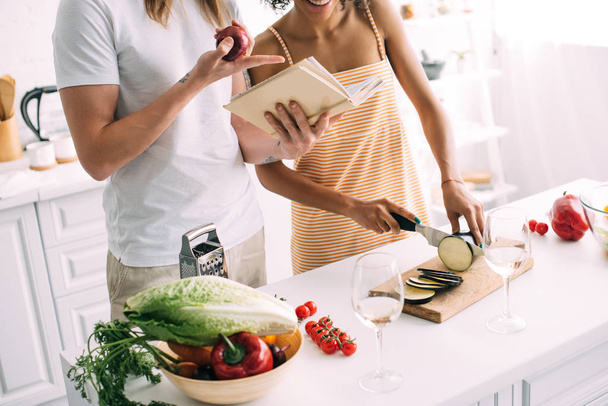 abgeschnittene Aufnahme eines Mannes mit Zwiebeln, der auf Rezept im Buch auf Freundin zeigt, die Auberginen schneidet  - Foto, Bild