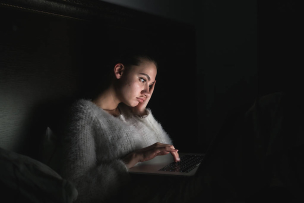 tired tired girl sitting in bed, in the dark, late at night, looking at laptop - 写真・画像
