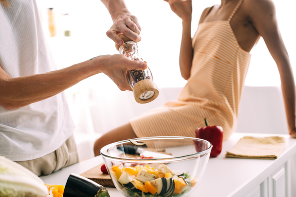 Oříznout záběr člověka pomocí mlýnek na pepř nad mísu se salátem a žena sedí na stole v kuchyni  - Fotografie, Obrázek