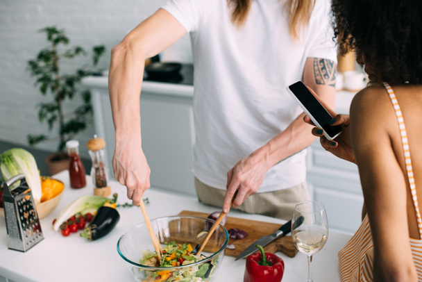 обрізане зображення чоловіка, який готує салат, а його дівчина стріляє на смартфон на кухні
  - Фото, зображення