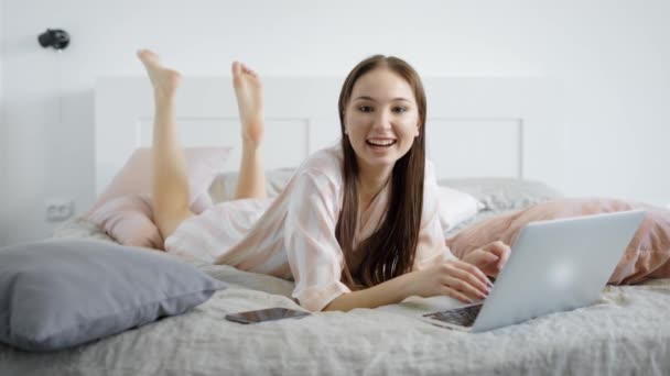 cheerful brown haired woman is lying on a bed in room in day with notebook in front of her and looking to a camera - Video
