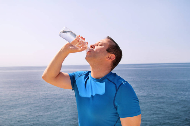 An athlete making a break during training, drinking fresh clean water from a bottle, rests and enjoys the clean air, with a beautiful landscape nature outdoors forest in background. Healthy lifestyle. - Photo, Image