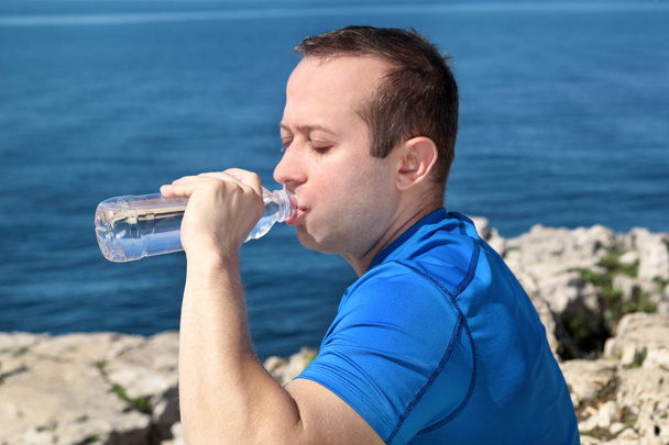 Un atleta haciendo una pausa durante el entrenamiento, bebiendo agua limpia fresca de una botella, descansa y disfruta del aire limpio, con una hermosa naturaleza paisajística al aire libre bosque en el fondo. Estilo de vida saludable
. - Foto, imagen