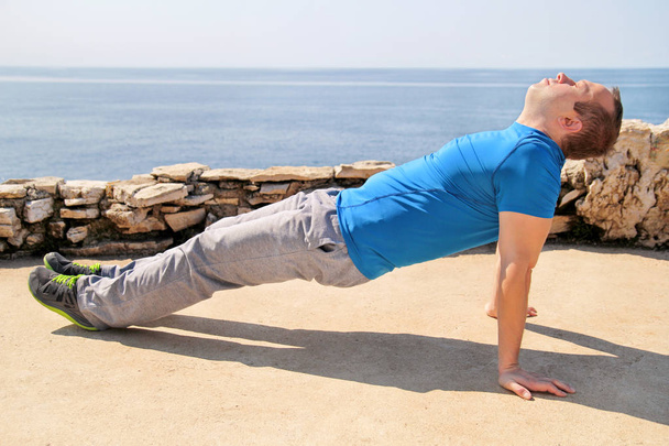 Een jonge man het beoefenen van yoga, pilates en stretching aan een kust. Knappe man doet uitrekkende oefeningen op het strand. Het beoefenen van een gezonde levensstijl. Fitness sporter doen yoga buiten. - Foto, afbeelding