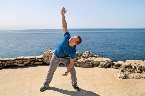 Athletic man doing stretching exercise, preparing for workout on seashore. Handsome fitness athlete doing a stretching routine, practicing yoga, meditation, pilates in sun. Sky and sea in background. - Photo, Image