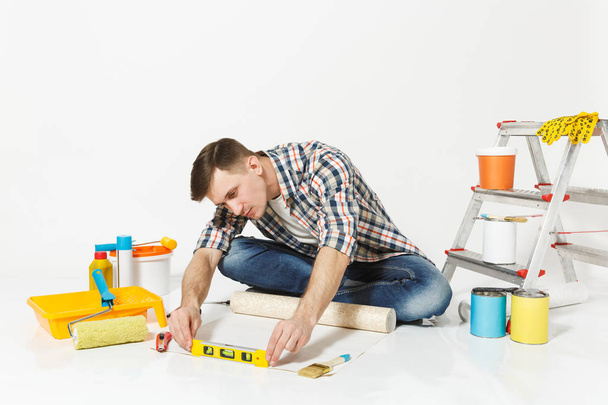 Młody człowiek siedzi na podłodze z rolki Tapety, budynek bąbelek poziomica, instrumenty na remont mieszkanie na białym tle. Klejenie akcesoria, narzędzi do malowania. Koncepcja domu naprawy - Zdjęcie, obraz