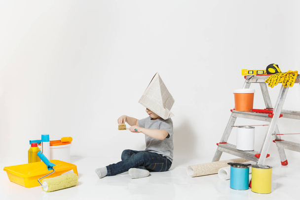Little cute boy in newspaper hat with instruments for renovation apartment room isolated on white background. Wallpaper, gluing accessories, painting tools. Repair home. Parenthood, childhood concept. - Фото, зображення