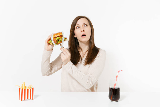 Frau am Tisch hält Burger mit Maßband herum, Pommes, Cola in Glasflasche isoliert auf weißem Hintergrund. richtige Ernährung oder klassisches amerikanisches Fast Food. Bereich mit Kopierraum. - Foto, Bild