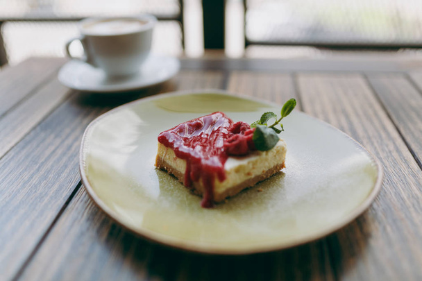 Lähikuva kakku vadelmakastiketta lautasella, kuppi cappuccino makaa puupöydällä kahvilassa kahvila ravintola. Yläpuolinen näkymä. Elämäntapakäsite. Kopioi avaruusmainos. Mainosalue
 - Valokuva, kuva